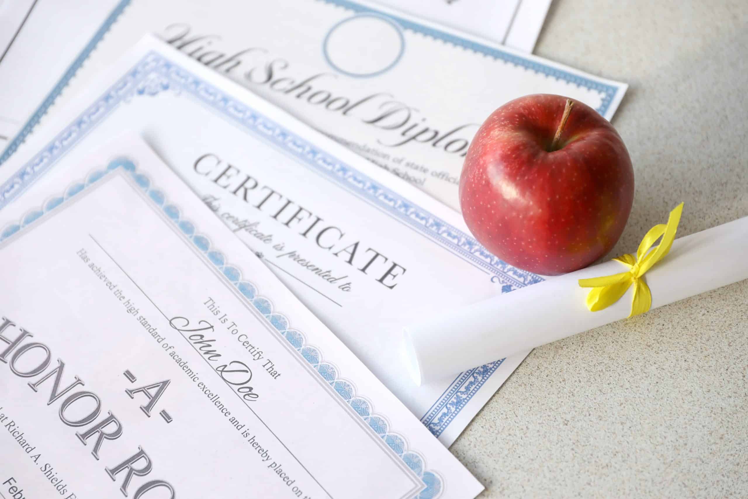a honor roll recognition, certificate of achievement and high school diploma lies on table with small scroll and red apple. education documents